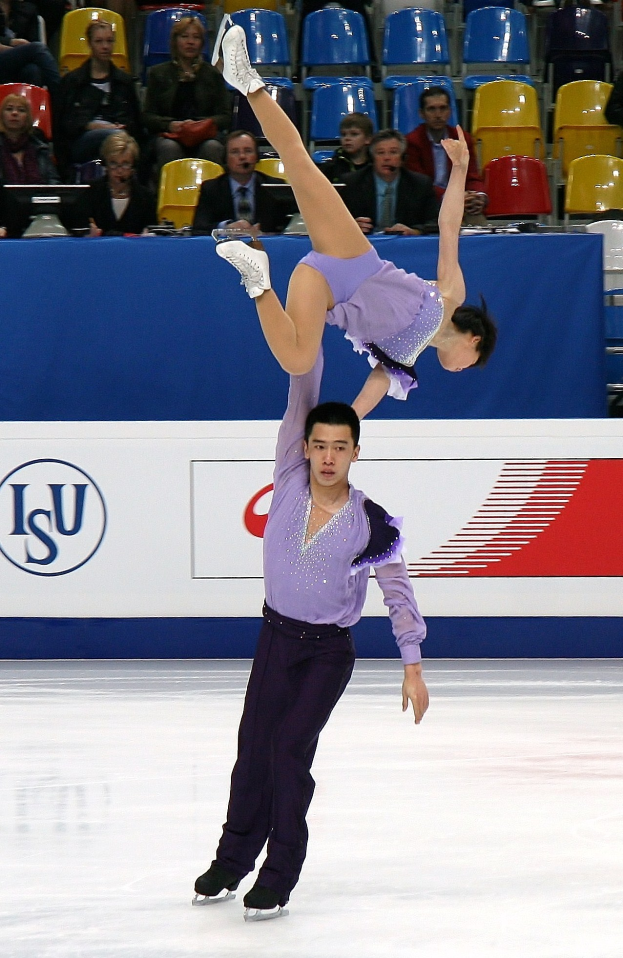 Zwei Eiskunstläufer, ein Mann in einem violetten Kleid und eine Frau in einem weißen Kleid, performen auf einer Eisbahn mit Zuschauern und einem blauen und weißen Schild im Hintergrund.