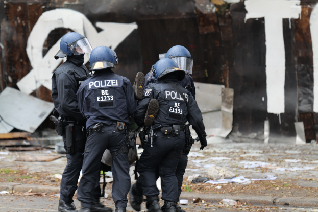 Polizeibeamte in schwarzen Uniformen und blauen Helmen stehen vor einem mit Graffiti bedeckten Gebäude während einer Ausschreitung, mit verstreuten Gegenständen und Text auf der Hintergrundwand.