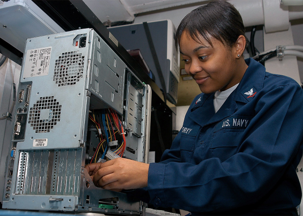 Eine Frau in einer blauen Uniform arbeitet an einem Computer in einem Raum mit elektronischen Geräten und Rohren an der Wand.