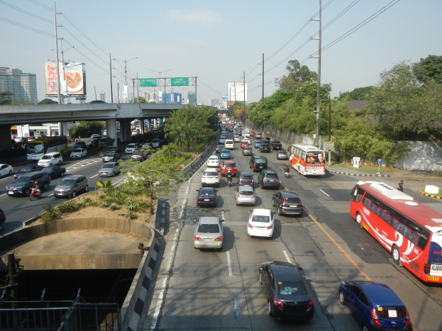 Eine belebte Stadtstraße mit Autos, Bussen, Strommasten, Bäumen, Gebäuden, Werbetafeln und einer Brücke unter einem bewölkten Himmel.