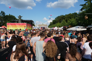 Eine große Menschenmenge geht eine Straße mit Zelten, Bäumen, Pfählen, Lichtern und einer Statue entlang, im Hintergrund sind Gebäude und ein Himmel mit Wolken und Ballons zu sehen bei der Christopher Street Day Parade in Berlin.
