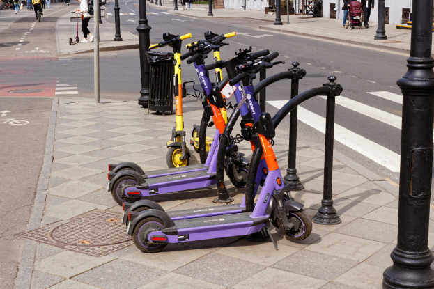 Eine Reihe von E-Scootern, die an einer Straße geparkt sind, mit Förderern, Radfahrern, einem Müllcontainer im Vordergrund und Gebäuden, Bäumen und Pflanzen im Hintergrund.