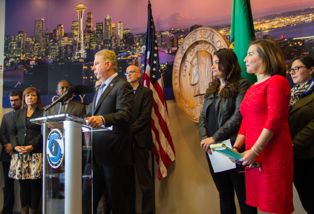 Ein Mann im Anzug spricht in ein Mikrofon an einem Rednerpult, mit einer Frau rechts daneben, vor einer Wand mit Flaggen und einer Tafel mit der Aufschrift "Seattle Mayor Bill de Blasio und die Stadt Seattle City Council".