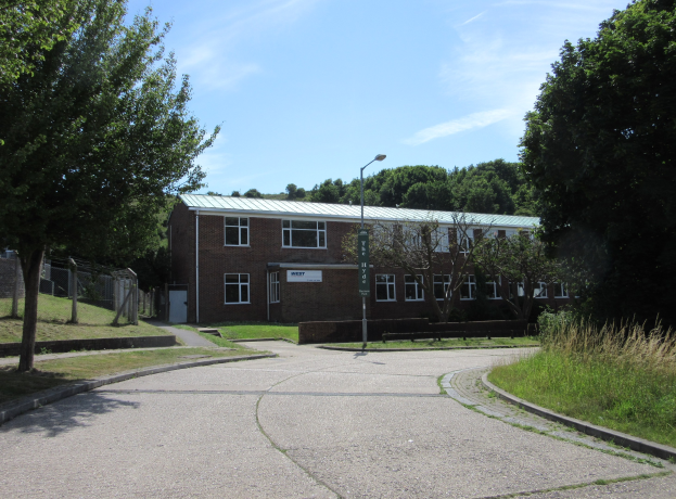 Ein Schulgebäude mit Fenstern, einer Straßenlaterne, einem Schild, einer Baumgruppe, Gras, einem Zaun und einem bewölkten Himmel, das in der Mitte eines Parks umgeben von saftigem Grün liegt.