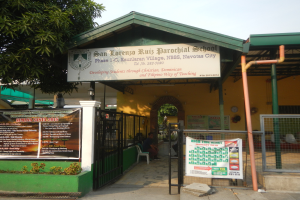 Der Eingang der San Lorenzo Ruiz Pfarrschule hat eine Tafel mit Text, einen Metallzaun, Menschen, die auf Stühlen unter einem Dach sitzen, Pfosten, ein Schild, Pflanzen, einen Baum und einen bewölkten Himmel.