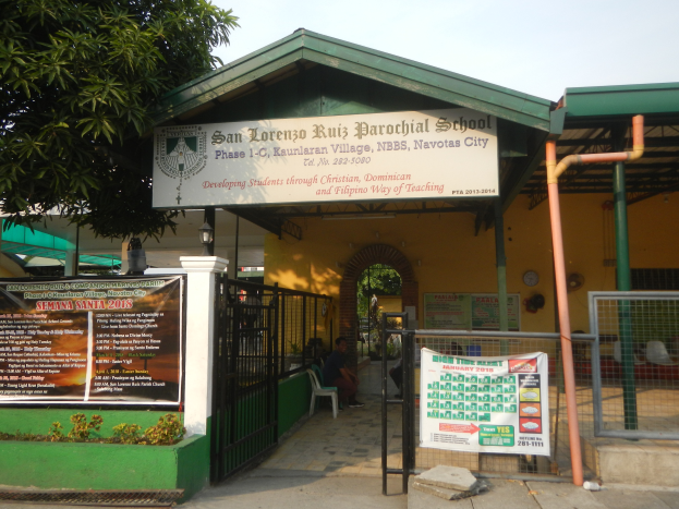 Der Eingang der San Lorenzo Ruiz Pfarrschule hat eine Tafel mit Text, einen Metallzaun, Menschen, die auf Stühlen unter einem Dach sitzen, Pfosten, ein Schild, Pflanzen, einen Baum und einen bewölkten Himmel.