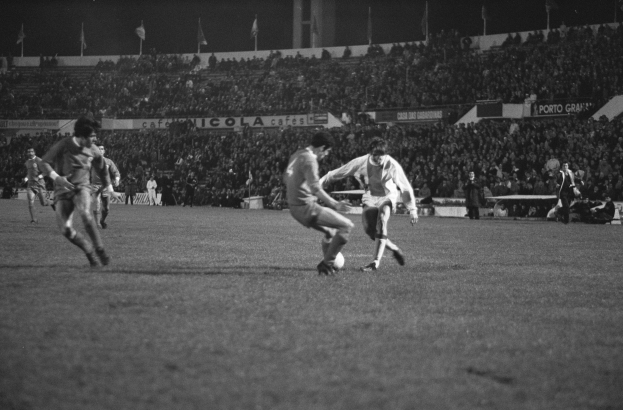 Schwarzes und weißes Foto von Männern, die Fußball auf einem Feld mit Zuschauern auf den Rängen und Fahnen im Hintergrund spielen.