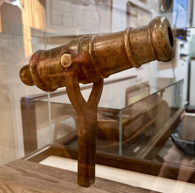 Alte Messingkanone in einer Glasvitrine auf einer hölzernen Oberfläche, mit gerahmten Gegenständen an der Wand im Hintergrund.
