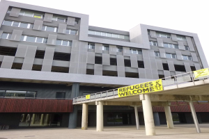 Großes Gebäude mit einem "Flüchtlinge Willkommen"-Schild, umgeben von Säulen und einem Geländer, vor einem Himmel-Hintergrund.