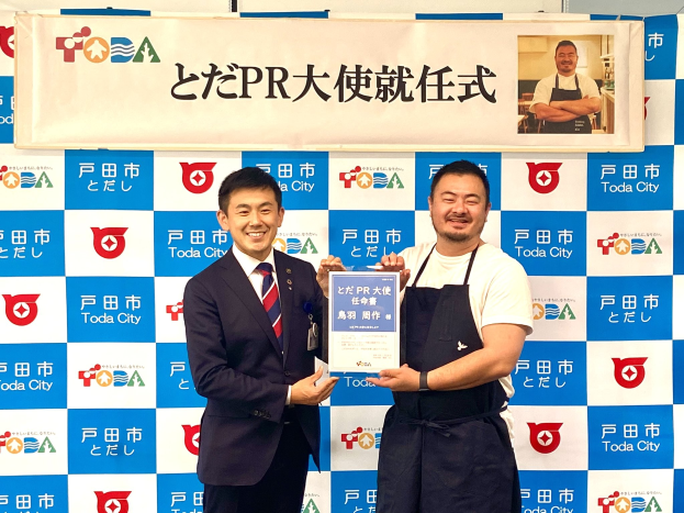 Zwei Männer mit einem Zertifikat, die lächeln, vor einem Banner mit der Aufschrift "Toda City Wins PR Award" und einem Foto im Hintergrund.