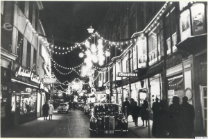 Eine Schwarz-Weiß-Aufnahme einer nächtlichen Stadtstraße mit fahrenden Autos, Fußgängern, Gebäuden mit erleuchteten Fenstern und Schildern sowie dekorativer Straßenbeleuchtung.