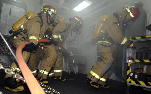Feuerwehrleute in Schutzbekleidung und Helmen arbeiten an einem Feuerlöschfahrzeug, mit Equipment-Racks und Hintergrundinfrastruktur im Hintergrund.