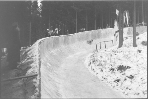 Ein Schwarz-Weiß-Foto einer Person, die auf einem Bobschlitten eine schneebedeckte Bahn hinunterfährt, umgeben von einem Zaun und Bäumen, mit ein paar Menschen im Hintergrund.