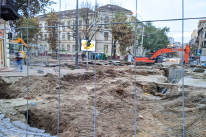 Baustelle mit einem ausgehobenen Bereich, der von einem Metallzaun umgeben ist, angrenzende Straße mit Fahrzeugen, Fußgänger auf einem Gehweg, Verkehrsampeln, Schilder, Bäume, Gebäude mit Fenstern und einen klaren blauen Himmel.