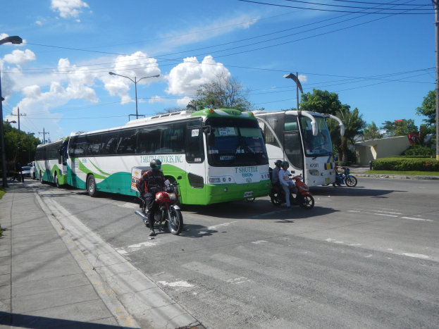 Ein grüner und weißer Shuttlebus parkt am Straßenrand mit Motorrädern davor, einem grasbewachsenen Fußweg links und Gebäuden, Bäumen und Laternenmasten im Hintergrund unter einem klaren blauen Himmel.