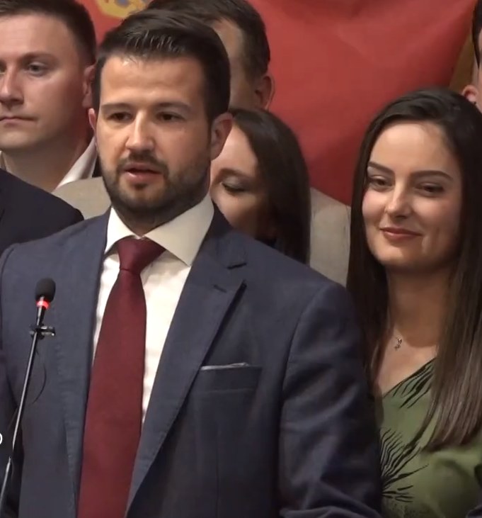 Mann im Anzug und Krawatte, der vor einem Publikum an einem Rednerpult steht und ein Mikrofon in der Hand hält, mit einem Banner im Hintergrund.