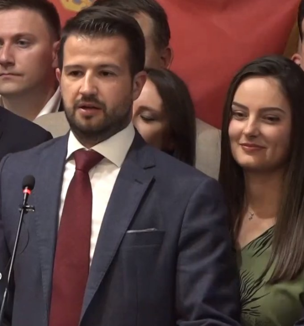 Mann im Anzug und Krawatte, der vor einem Publikum an einem Rednerpult steht und ein Mikrofon in der Hand hält, mit einem Banner im Hintergrund.