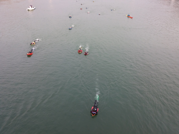 Gruppe von Menschen, die auf einem See mit Bäumen im Hintergrund Kajaks paddeln, während sie an einem Wassersport-Event teilnehmen.