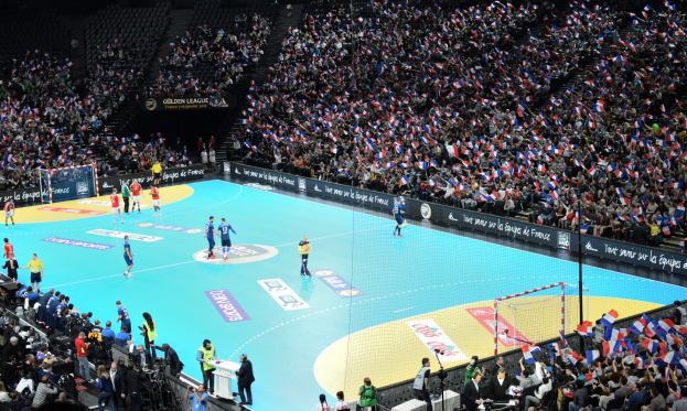 Eine Gruppe von Menschen spielt Handball in einer Arena mit einem Netz, das das Spielfeld teilt, und Zuschauern auf den Tribünen, während Fotografen die Aktion aufnehmen.