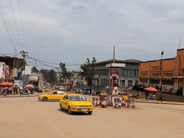 Ein gelbes Taxi fährt eine belebte Stadtstraße entlang, die von Gebäuden gesäumt ist, während Fußgänger auf dem Gehweg gehen, einige halten Schirme, unter einem bewölkten Himmel mit sichtbaren Strommasten und Kabeln.