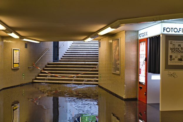 Überschwemmte U-Bahn-Station in Berlin mit Wasser auf dem Boden, Treppen mit Geländern, gerahmte Fotos an den Wänden, Deckenlampen und einen Getränkeautomaten.