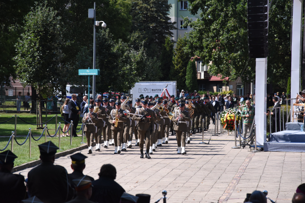 Eine Gruppe von Menschen in Möppen und mit Musikinstrumenten marschiert durch eine straßenlinke Straße, mit einem Podium mit Mikrofon und Ständer im Vordergrund und einer Zuschauermenge.