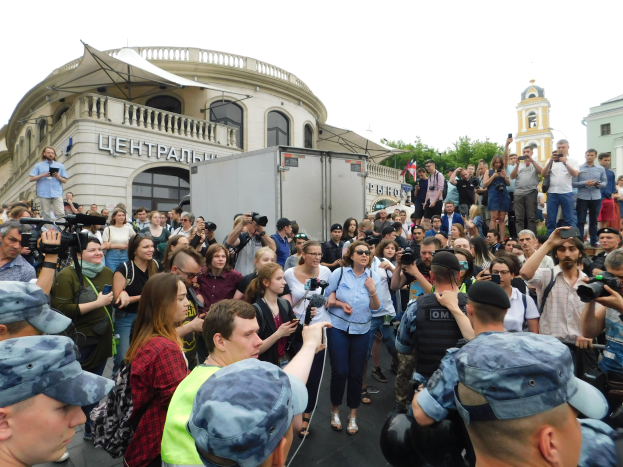 Große Menschenmenge vor einem Gebäude protestierend, einige halten Kameras und Handys, mit einem Fahrzeug, Bäumen und klarem blauem Himmel im Hintergrund.