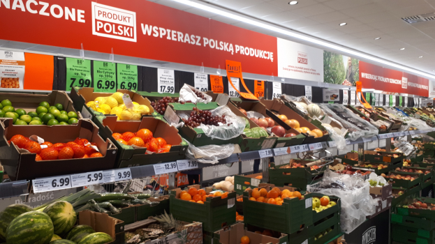 Ladeninneres mit verschiedenen Obst- und Gemüsesorten in Kartons mit Preisschildern, Wandtafeln mit Text und Bildern sowie Deckenleuchten.