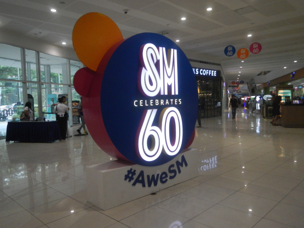 Eine Gruppe von Menschen steht in einem Einkaufszentrum mit einem Schild, auf dem "SM feiert 60-jähriges Jubiläum" steht, in der Mitte, einem Tisch mit schwarzem Tuch und Gegenständen links daneben und Glasfenstern und Schildern im Hintergrund.