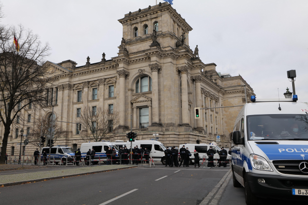 Eine Gruppe von Polizisten steht vor dem Reichstaggebäude in Berlin, Deutschland, mit Fahrzeugen, einem Zaun, Verkehrszeichen, Laternen, Bäumen und Flaggen im Hintergrund, unter einem klaren Himmel.