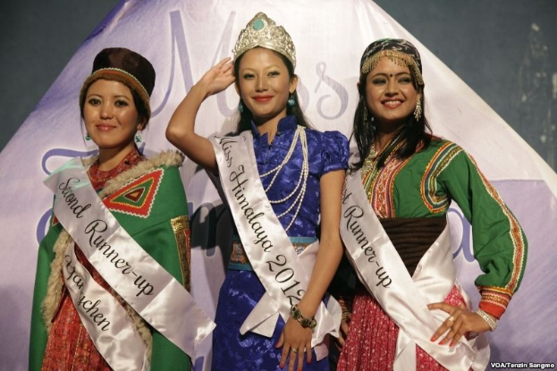 Drei Frauen in Schärpen und Kronen stehen zusammen auf einer Bühne mit einem Banner und Text, der unten 'Miss Himalaya 2012' liest.