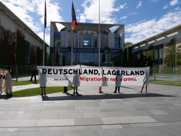 Eine Gruppe von Menschen mit einem Transparent mit der Aufschrift "Deutschland, Lagerland Migration ist kein Verbrechen" vor einem umzäunten Gebäude mit Bäumen und Flaggen.