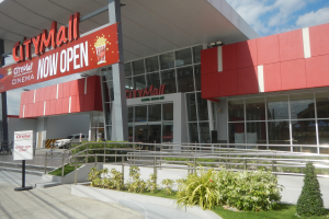 Ein Stadtzentrum mit einem Schild, auf dem "Stadtzentrum jetzt offen" steht, umgeben von Pflanzen, Gras, einem Metallzaun, Menschen, einem Auto, Pfosten, einer Fahne, Lampen und einem bewölkten Himmel.