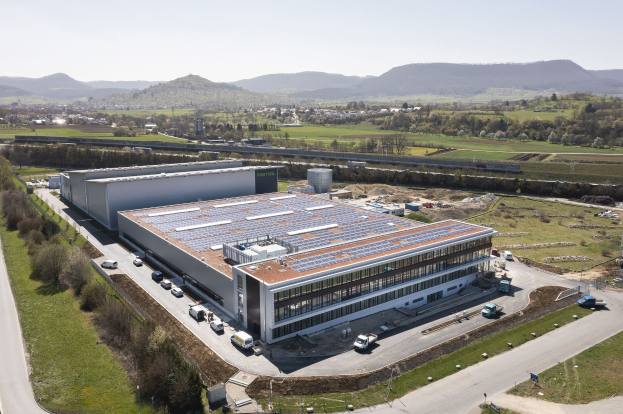 Luftaufnahme eines großen Gebäudes mit Solarmodulen auf dem Dach, umgeben von Fahrzeugen, Bäumen, Gras, Pfählen und Hügeln unter einem klaren blauen Himmel, identifiziert als Deutschlands erste solarbetriebene Fabrik.