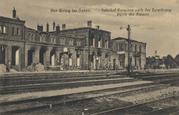 Schwarzes und weißes Foto des Bahnhofs Korschen in Ostern, Deutschland, das Gleise, Bahnhofsgebäude, Menschen, Bäume und Himmel zeigt.