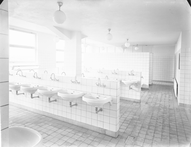 Schwarz-weißes Foto einer öffentlichen Toilette mit Waschbecken, Wasserhähnen, Wänden, Lampen und Fenstern.