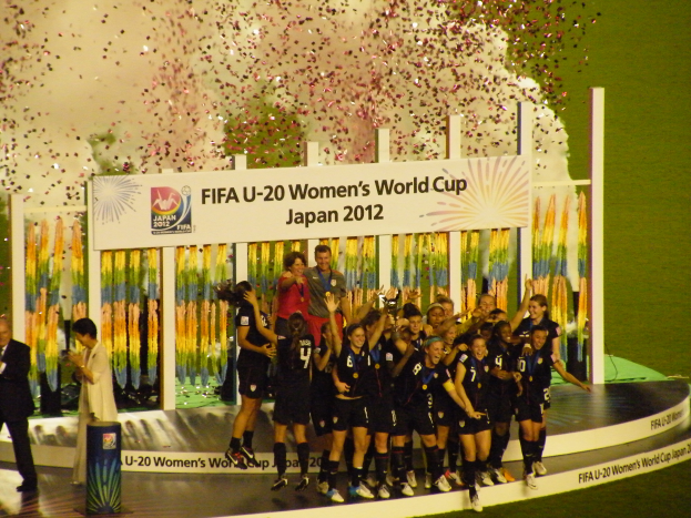Eine Gruppe von Fussballspielerinnen in schwarzen Uniformen steht auf einer Bühne, feiert ihren Sieg und ist von einer Menge mit Kameras umgeben, mit einem dekorierten Hintergrund und grasbewachsenem Boden.