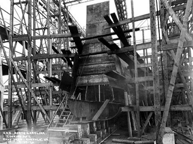 Schwarzes und weißes Foto eines Schiffes in einem Schiffbau, mit hölzernen Pfählen und Treppen im Vordergrund und Himmel im Hintergrund, mit Text "Marinebewaffnung" in der linken unteren Ecke.