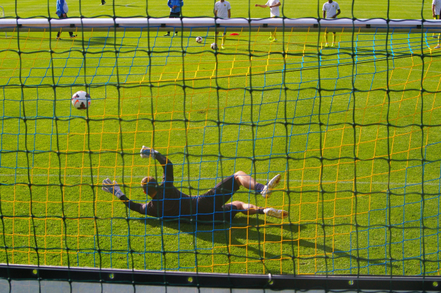 Ein Fussballer hält den Ball vor einem Tor, mit einem Netz im Vordergrund und einigen Personen im Hintergrund sowie mehreren Bällen auf dem Boden verstreut.