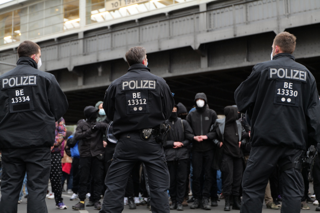 Polizeibeamte in schwarzen Uniformen und Masken vor einer Menge mit einer Brücke und einem Gebäude im Hintergrund während einer Demonstration.