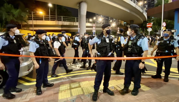 Polizisten in Uniform und Masken stehen vor einer Menge, mit Fahrzeugen, Gebäuden, Bäumen und einer Brücke im Hintergrund, wahrscheinlich nach einer Demonstration in Hong Kong.