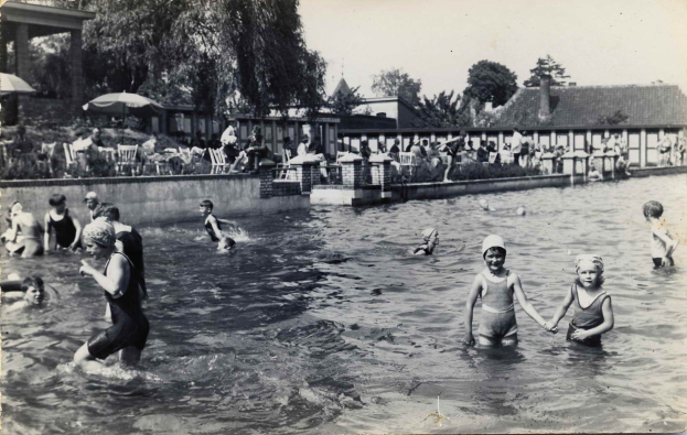 Schwarzes und weißes Foto von Kindern, die in einem Swimmingpool spielen, mit Menschen auf Stühlen und Schirmen im Hintergrund.
