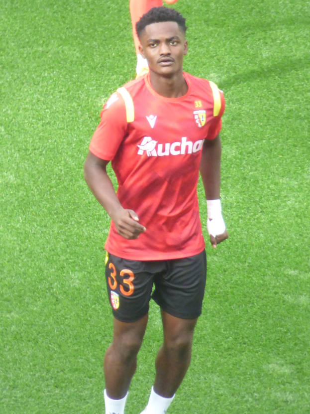 Emmanuel Adebayor, der ein rotes T-Shirt und schwarze Shorts trägt, geht auf einem Fussballfeld mit zwei Personen im Hintergrund.
