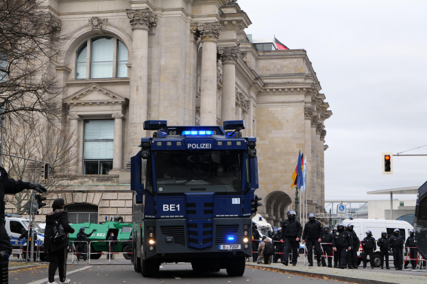 Eine Gruppe von Polizeibeamten steht vor einem großen Gebäude mit Fahrzeugen auf der Straße und einer Person, die eine Kamera hält, links daneben, mit Bäumen, Verkehrszeichen, Fahnen und einem klaren blauen Himmel im Hintergrund.