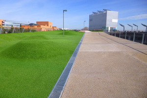 Grünes Dach mit Kunstgras, umgeben von Gebäuden, Straßenlaternen, Gittern, Bäumen und einem bewölkten Himmel.