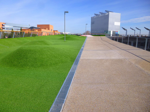 Grünes Dach mit Kunstgras, umgeben von Gebäuden, Straßenlaternen, Gittern, Bäumen und einem bewölkten Himmel.
