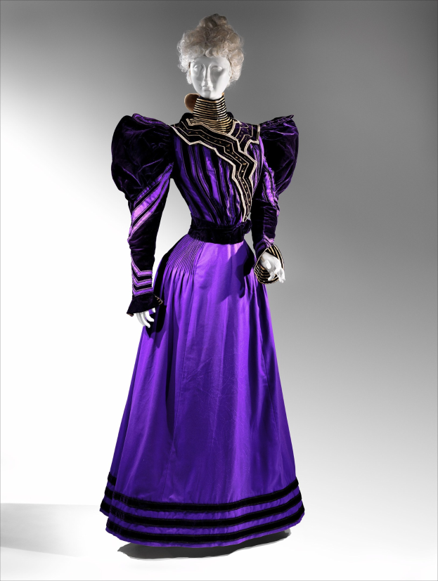 Eine Schaufensterpuppe in einem lila Abendkleid mit schwarzem und goldenem Muster, das eine eng anliegende Taille, einen langen Rock bis zum Boden mit goldener Stickerei und Perlen sowie eine Spitzenborte an der Taille hat, kombiniert mit einem eleganten Hairstyle und zarten Ohrringen.