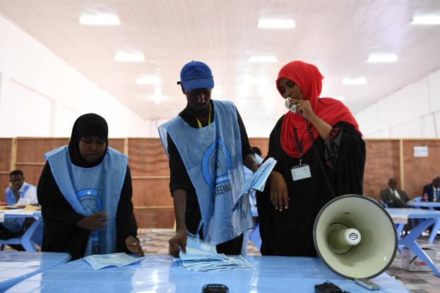 Menschen versammeln sich um einen Tisch mit einem Megafon und Papieren, die scheinbar eine Wahllokalität darstellen, während andere im Hintergrund sitzen und stehen, unter beleuchteten Decken.