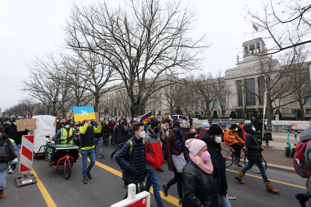 Eine große Gruppe von Menschen marschiert auf einer Demonstration in Washington, D.C. am 21. Januar 2020 mit Schildern, Bannern und Fahrrädern, vor einem Gebäude und Bäumen unter einem klaren blauen Himmel.