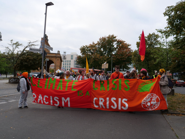 Gruppe von Menschen marschiert eine von Bäumen gesäumte Straße entlang und hält ein 'Klimakrise ist eine Krise'-Schild, mit Fahrzeugen, Gebäuden und einem klaren blauen Himmel im Hintergrund.